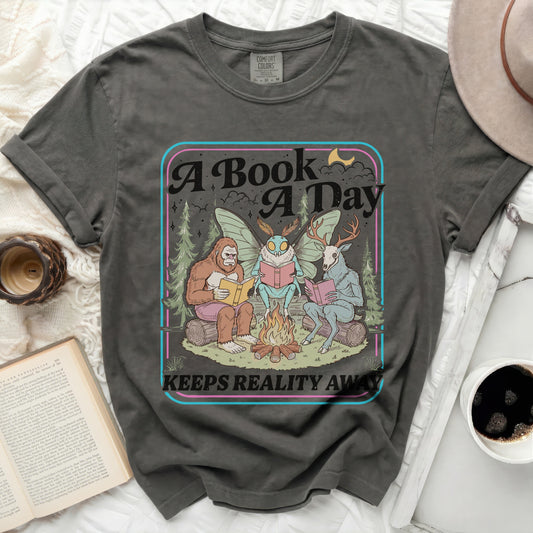 Dark grey t-shirt with Bigfoot, moth, and deer reading by a campfire.