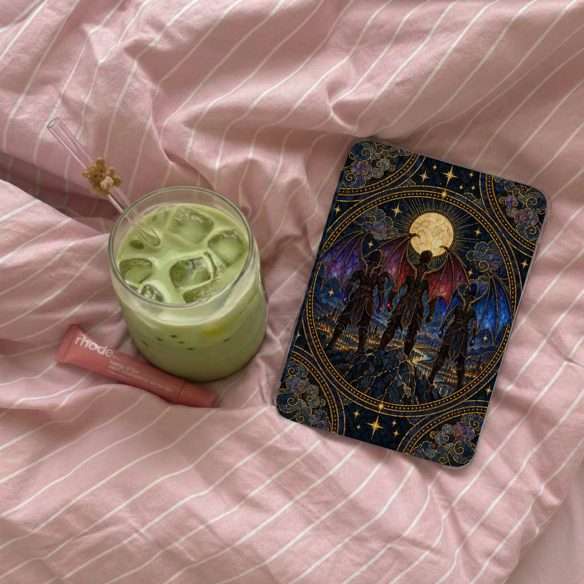 An e-reader with an ornate fantasy art case rests on pink striped bedding next to an iced matcha latte and lip balm.
