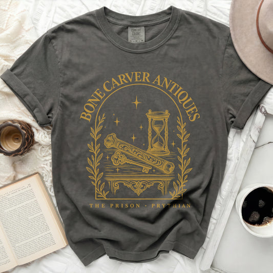 A dark gray crewneck t-shirt featuring a gold print with an hourglass, key, bone, and text reading "Bone Carver Antiques The Prison Prythian."