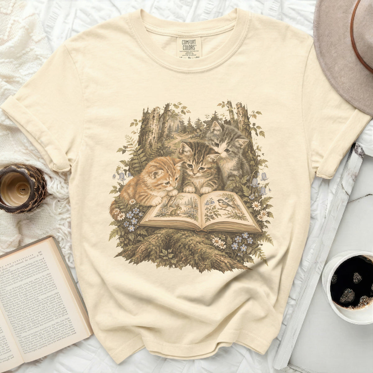 Cream t-shirt with a vintage illustration of three kittens reading a book in a forest.