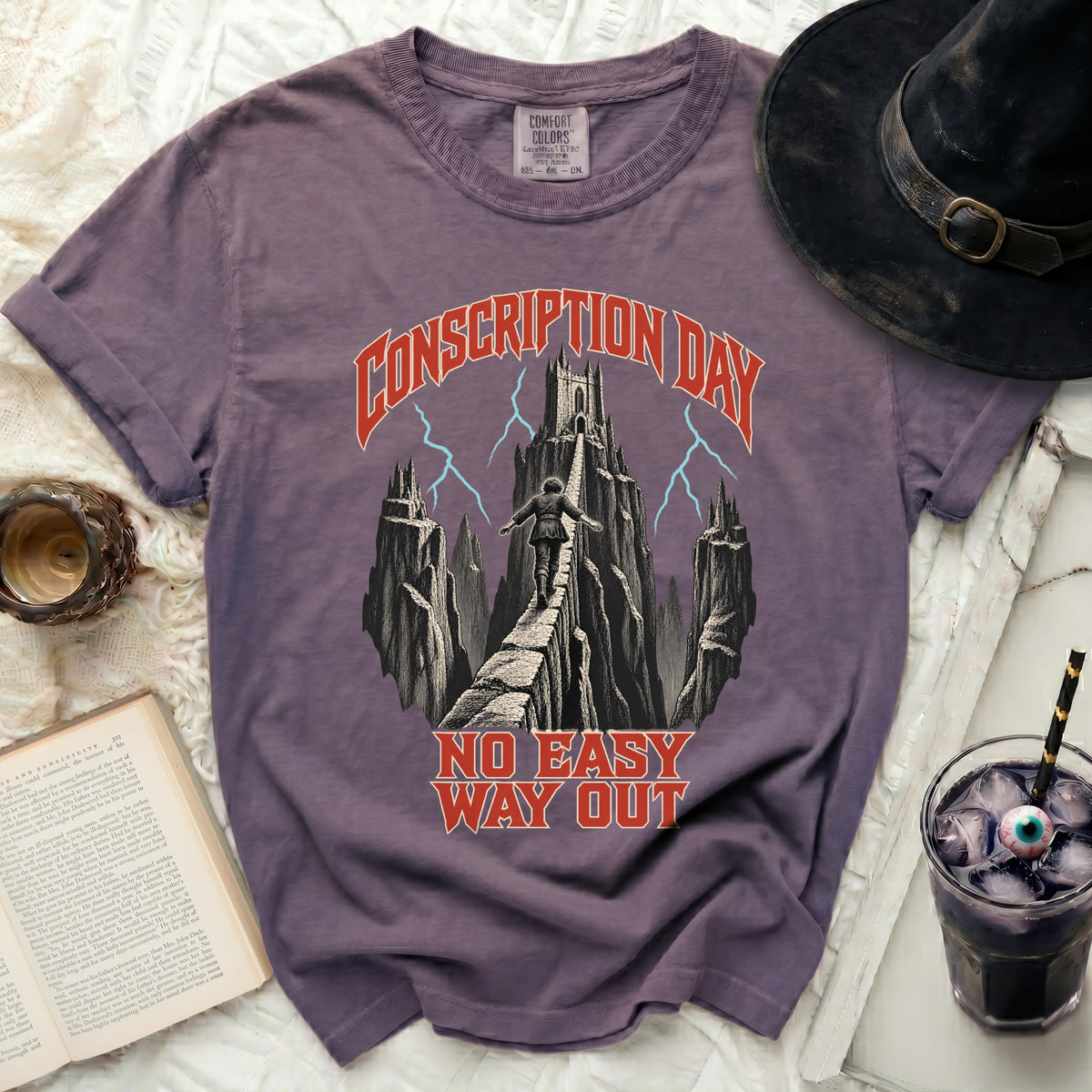 A purple t-shirt featuring a Conscription Day graphic with text and a figure crossing a mountain bridge.