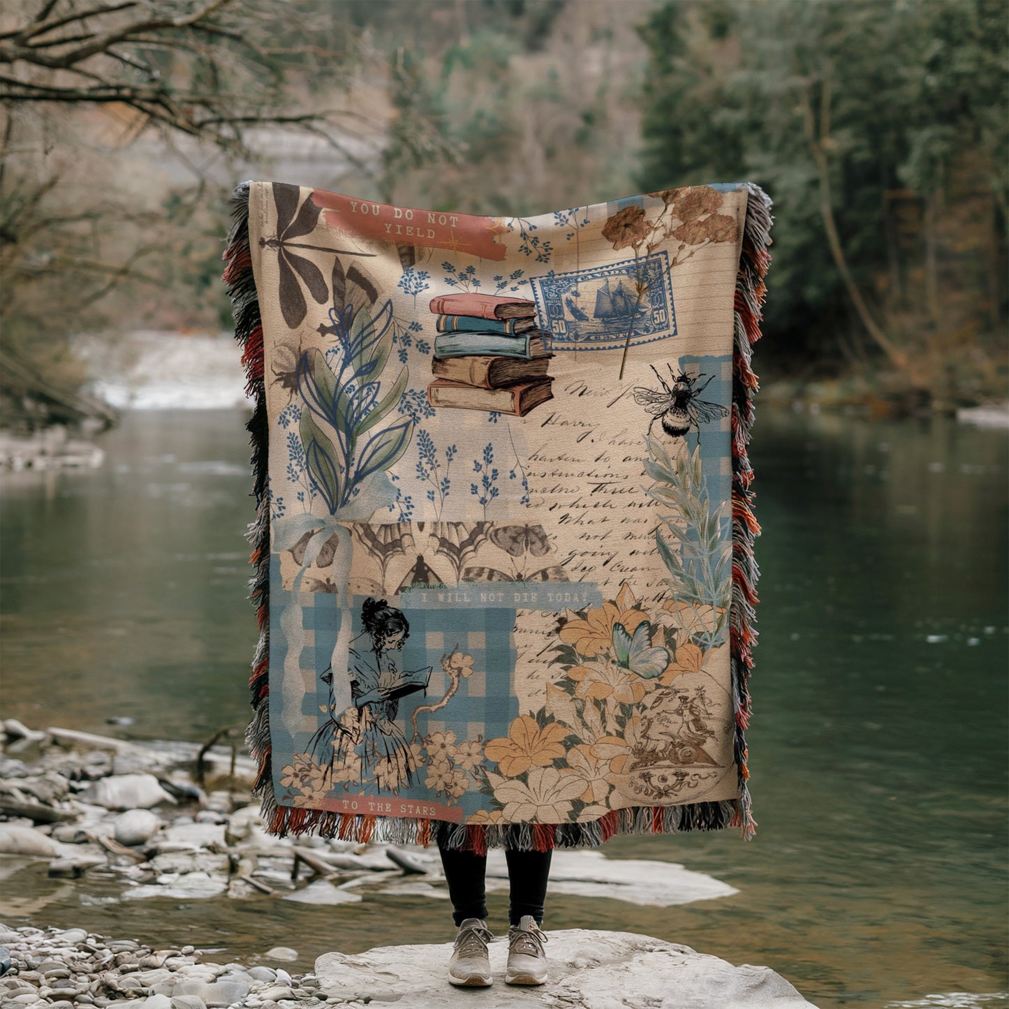 A person stands on a rock, holding a colorful blanket with various illustrations and text, including a book and a bee, in front of a body of water.