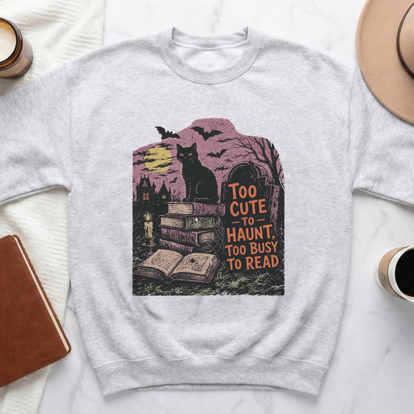 Grey sweatshirt with black cat, books, gravestone, and text "Too cute to haunt, too busy to read."
