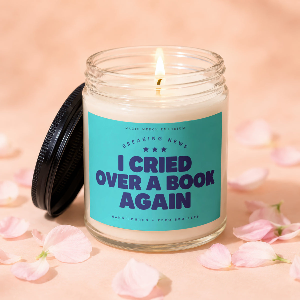 A lit candle in a glass jar with a turquoise label reading "I Cried Over A Book Again," surrounded by pink flower petals.