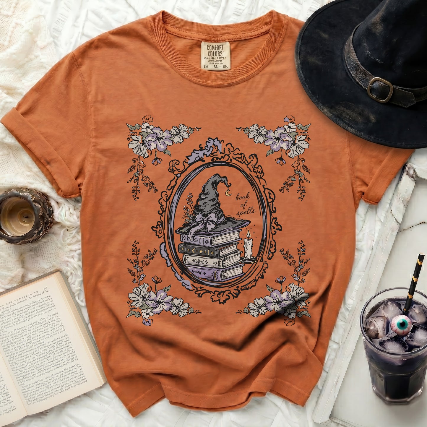 Orange t-shirt with a vintage-style graphic of a witch's hat, books, and flowers.