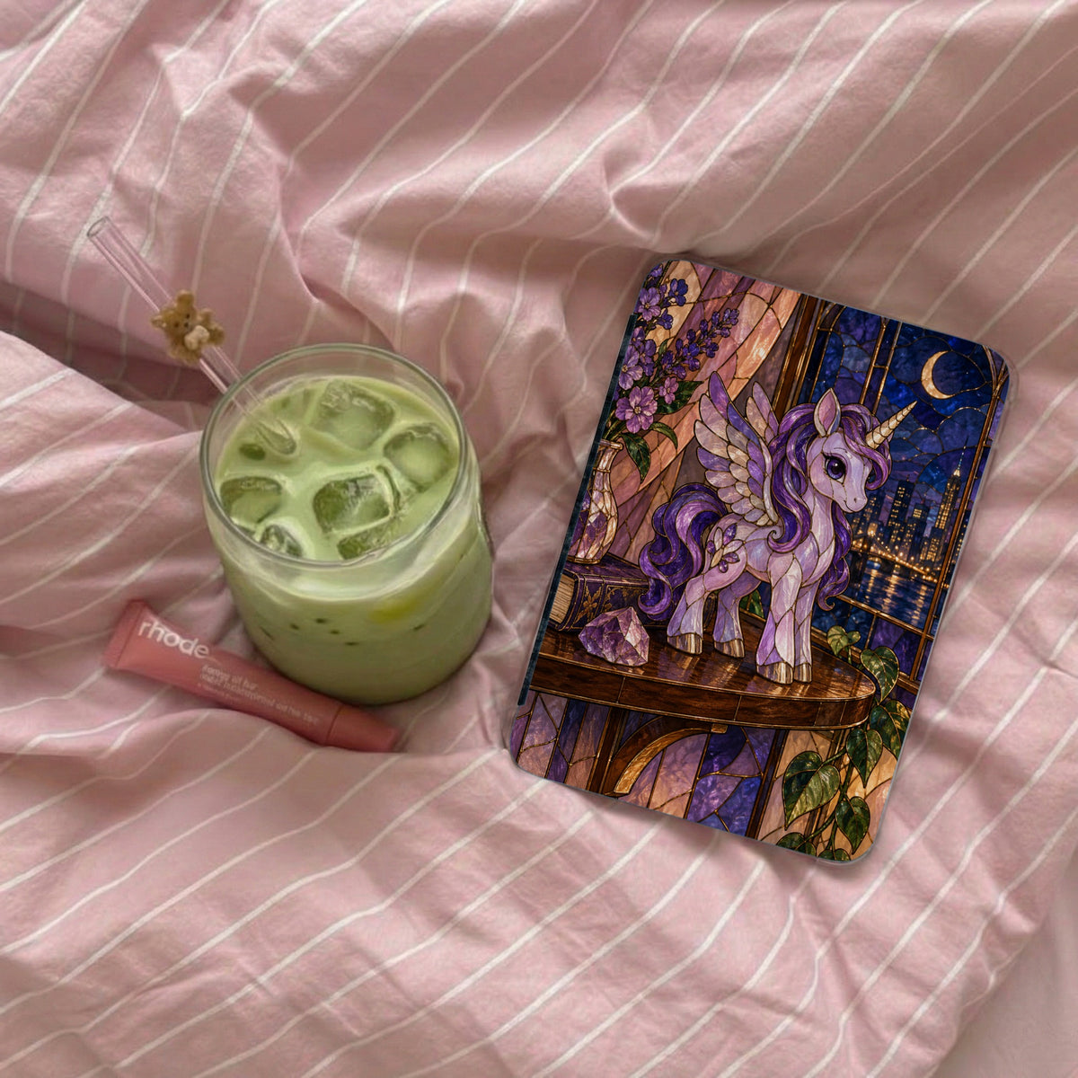 Stained glass unicorn journal with cityscape, matcha drink, and lip balm on pink striped fabric.