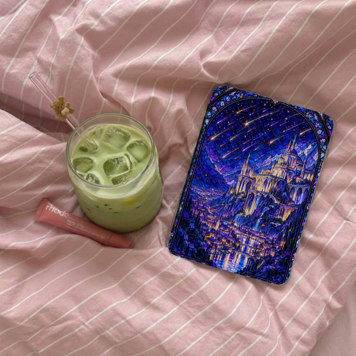 A blue fantasy castle tablet case rests on a pink striped bedsheet beside an iced matcha latte.