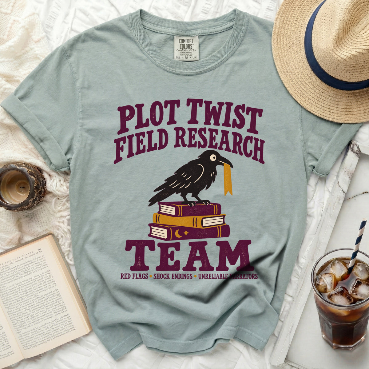 Light blue t-shirt with "Plot Twist Field Research Team" text, crow on books.