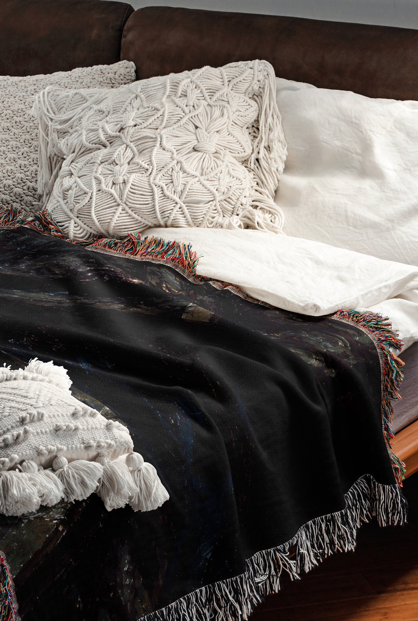 a cozy and inviting bedroom scene, featuring a bed with a black blanket and white pillows, a black throw blanket with fringe, and a white throw blanket with fringe. The bed is positioned against a brown leather couch, creating a comfortable and stylish living space.