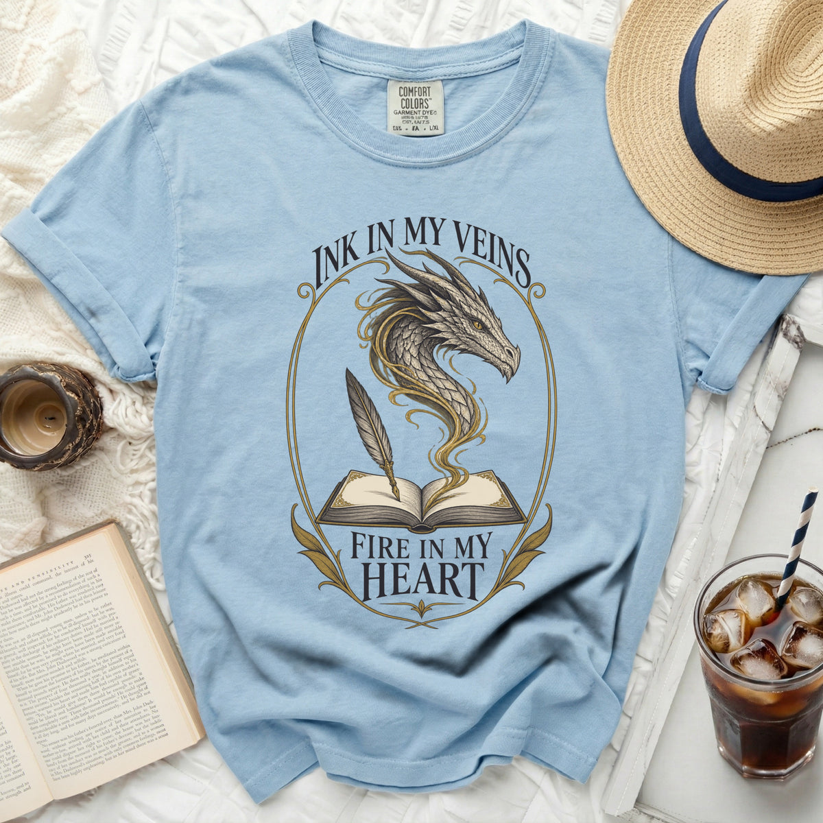 Light blue t-shirt with dragon, book, and feather graphic. Text reads "Ink in my veins, fire in my heart."