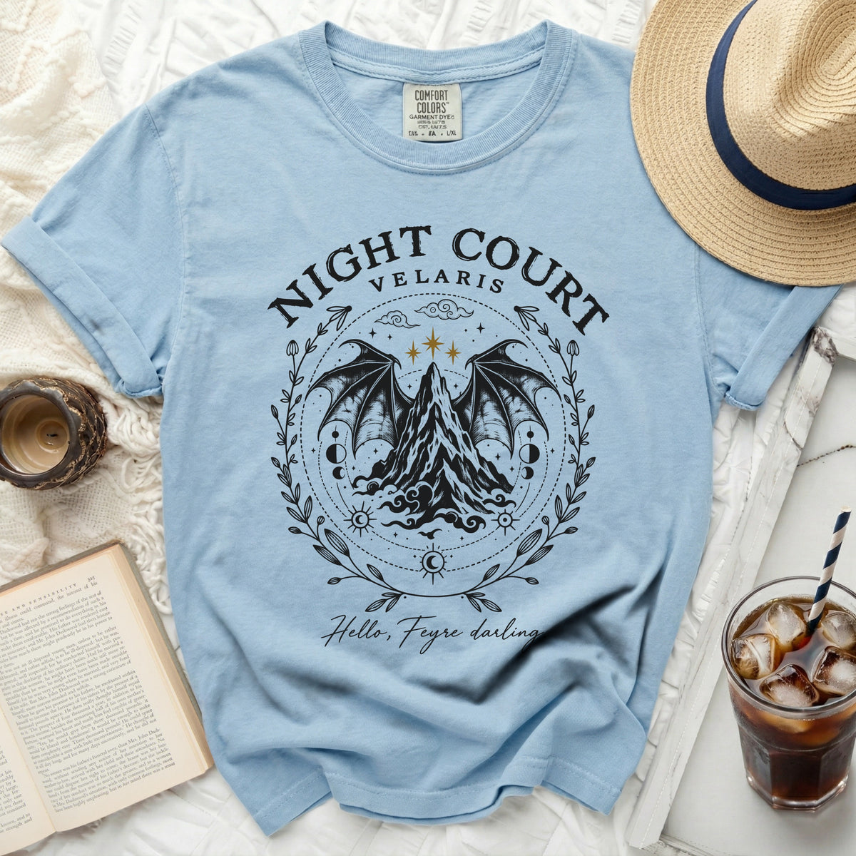 A light blue t-shirt featuring a Night Court Velaris graphic, surrounded by books and a drink.