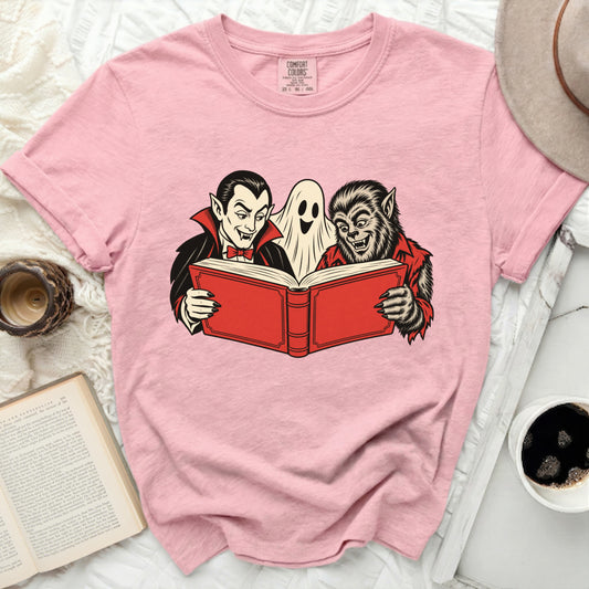 Pink t-shirt with graphic of vampire, ghost, and werewolf reading a book.