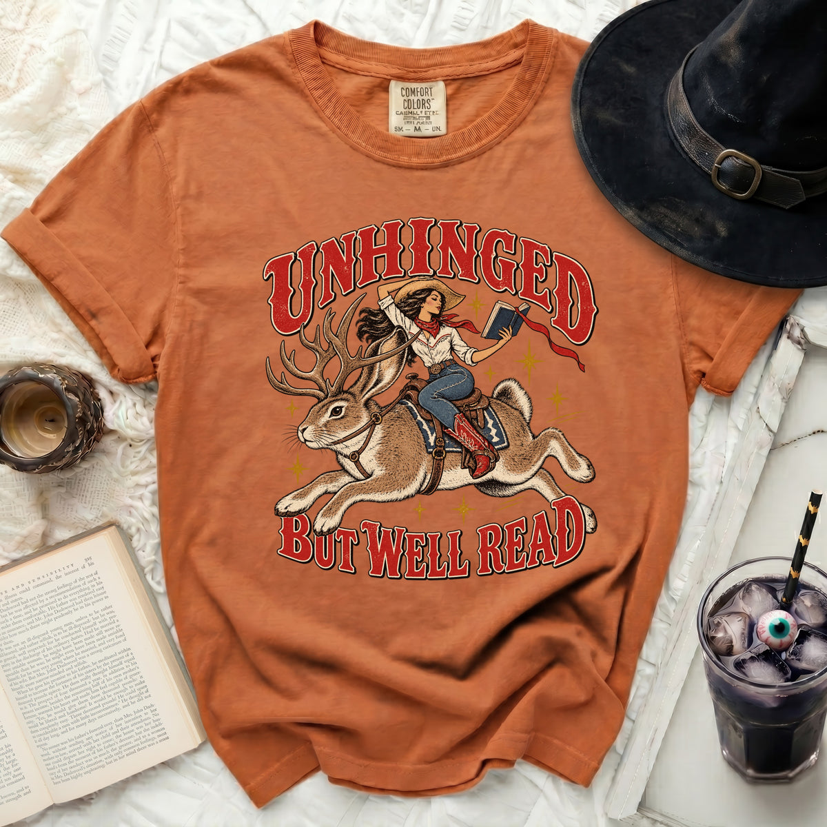 Orange t-shirt with "Unhinged But Well Read" graphic of cowgirl riding jackalope.