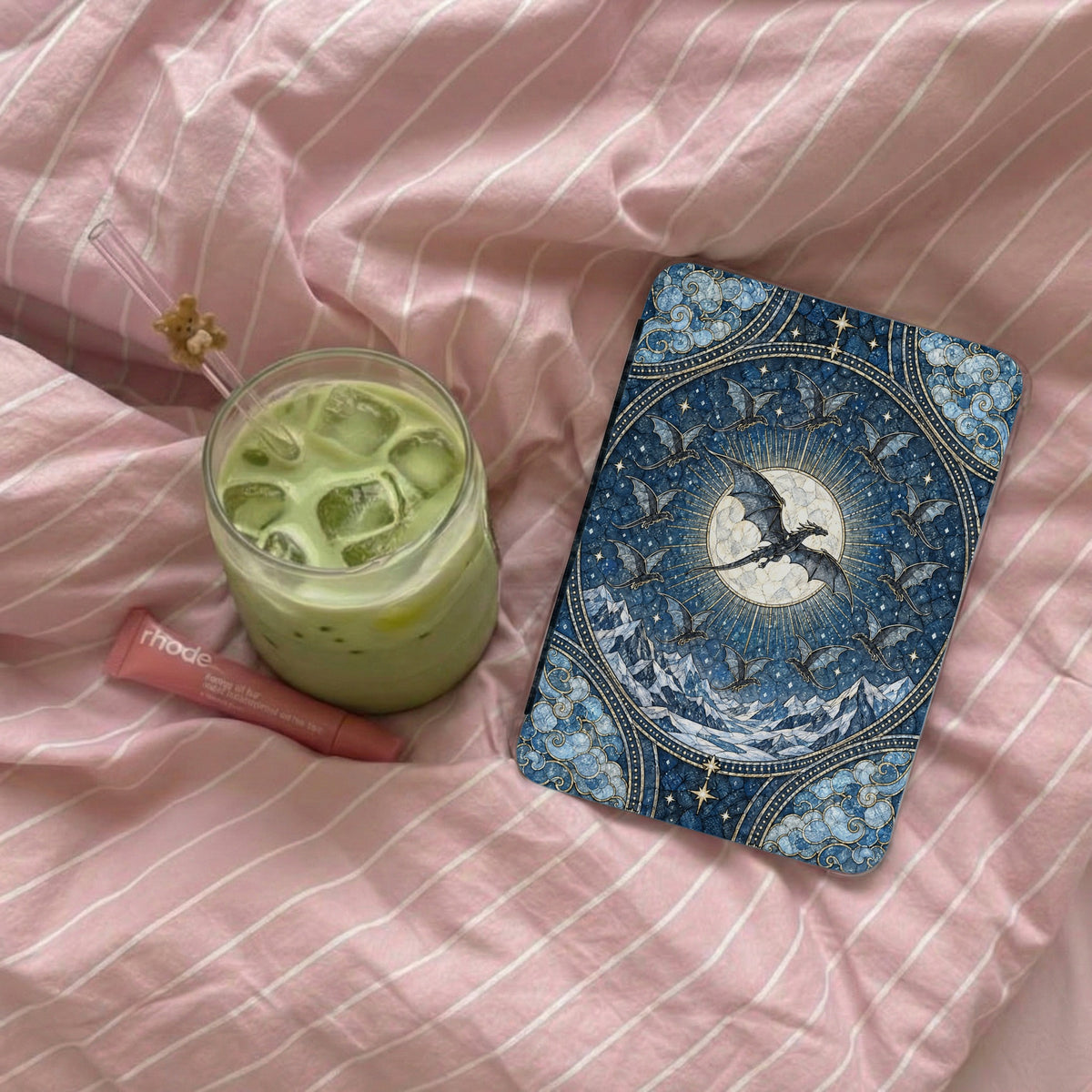 An e-reader with an ornate blue dragon cover rests on a pink-striped comforter beside a matcha iced latte.
