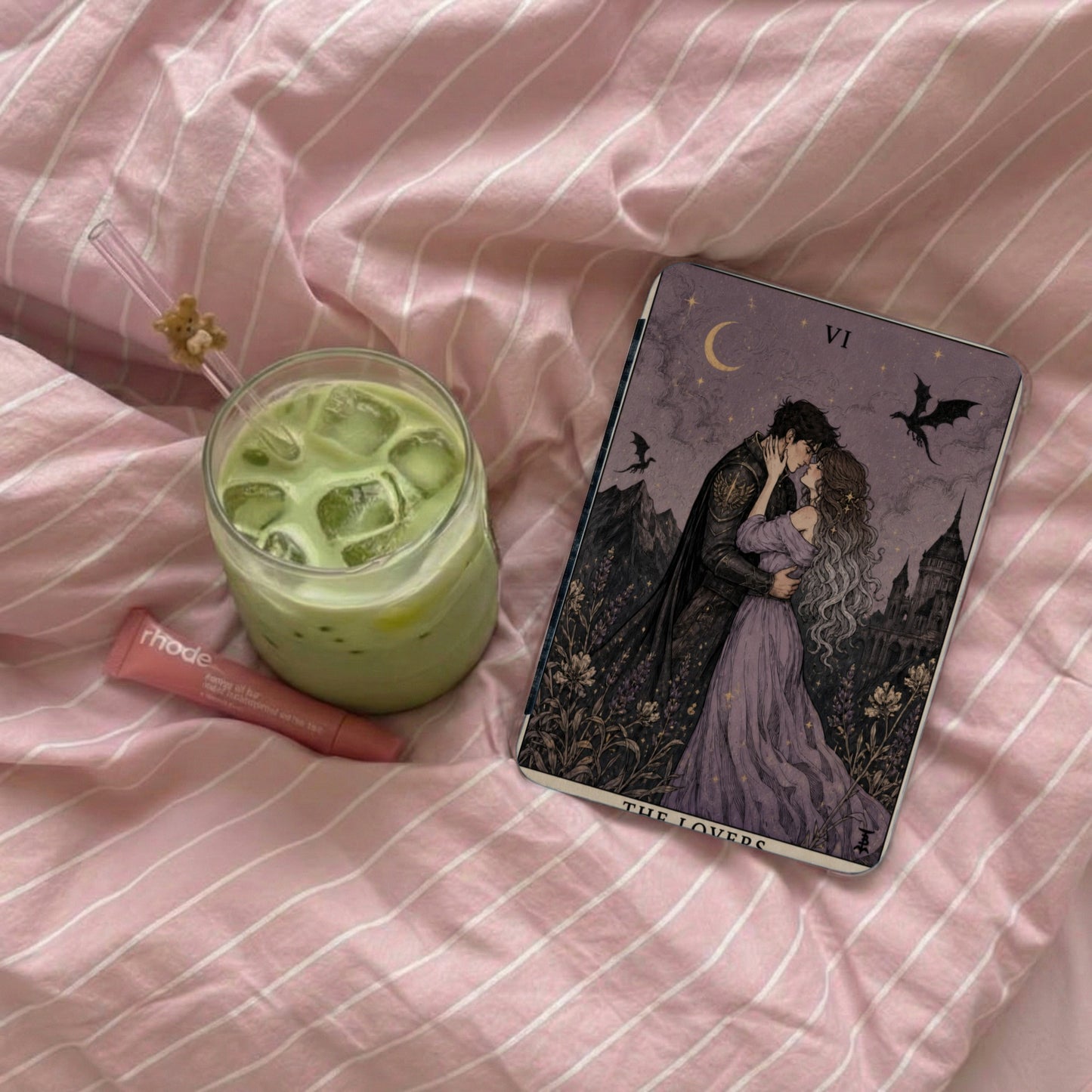 "The Lovers" tarot card with a matcha drink and lip balm on pink striped bedding.