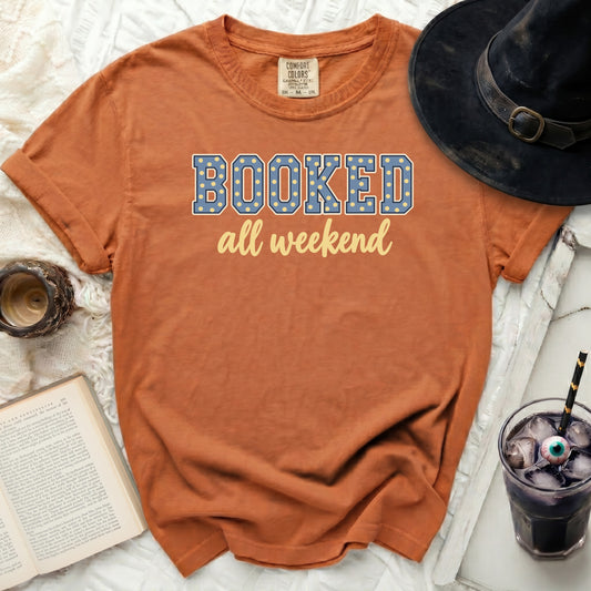 Orange t-shirt with "BOOKED all weekend" text, witch hat, book, and drink.