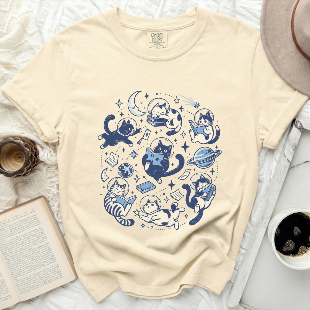 Cream t-shirt with blue astronaut cats reading books and floating in space.