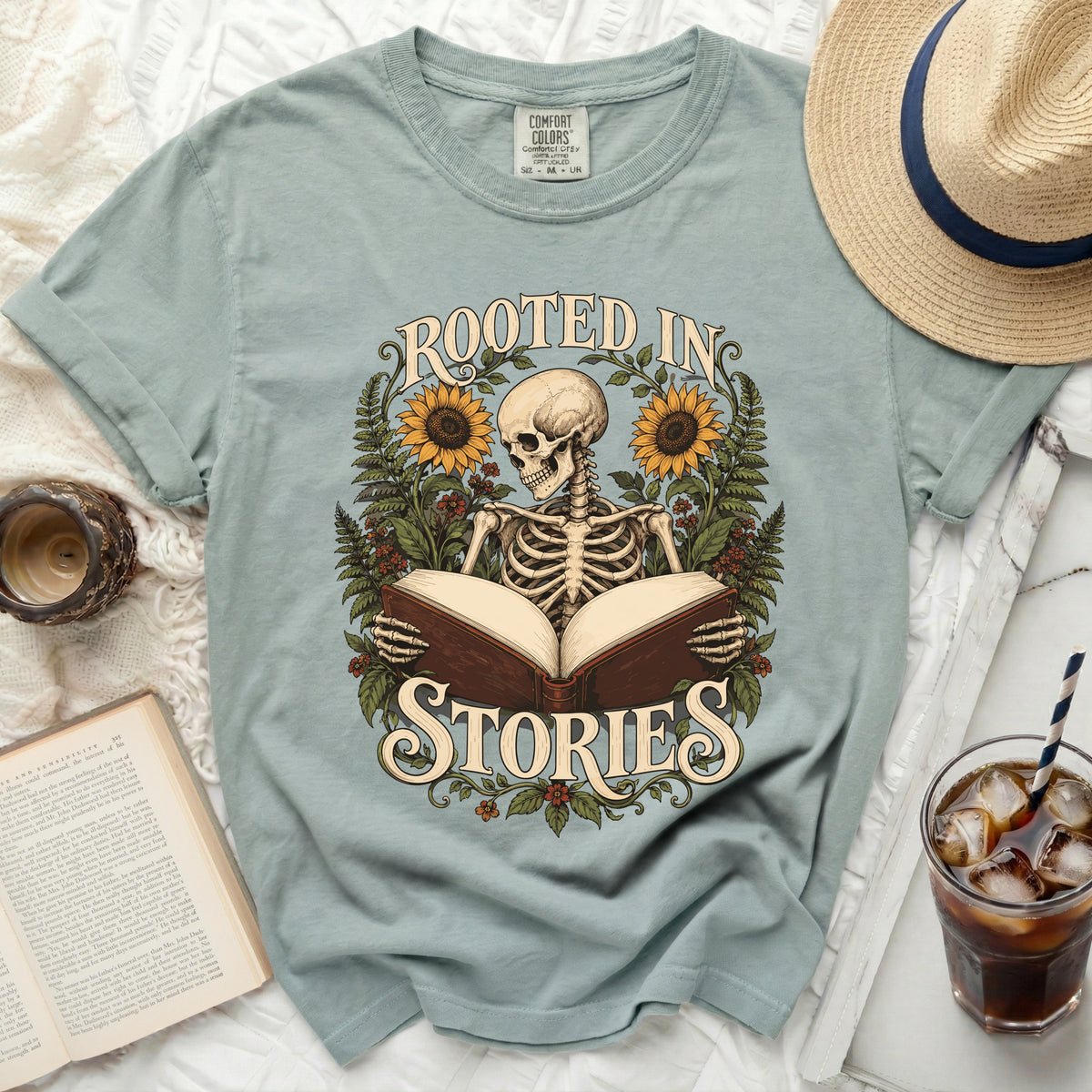 Skeleton reading a book with sunflowers and text "ROOTED IN STORIES" on t-shirt.