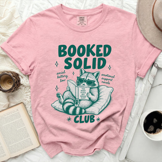 Pink t-shirt with raccoon reading "Too Many Plots, Not Enough Sleep" and "Booked Solid Club" text.