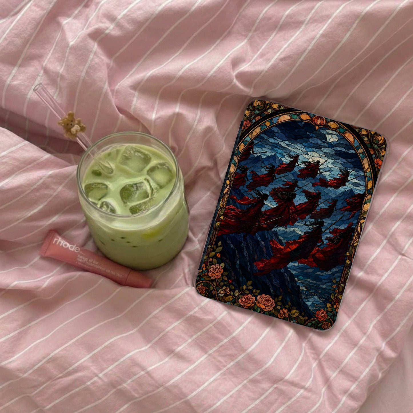 Glass of iced matcha with a straw, pink lip balm, and a stained glass art piece.