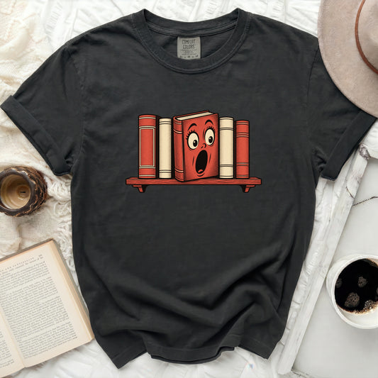 A graphic t-shirt featuring a distressed dark gray fabric with a central illustration of cartoon books on a shelf.