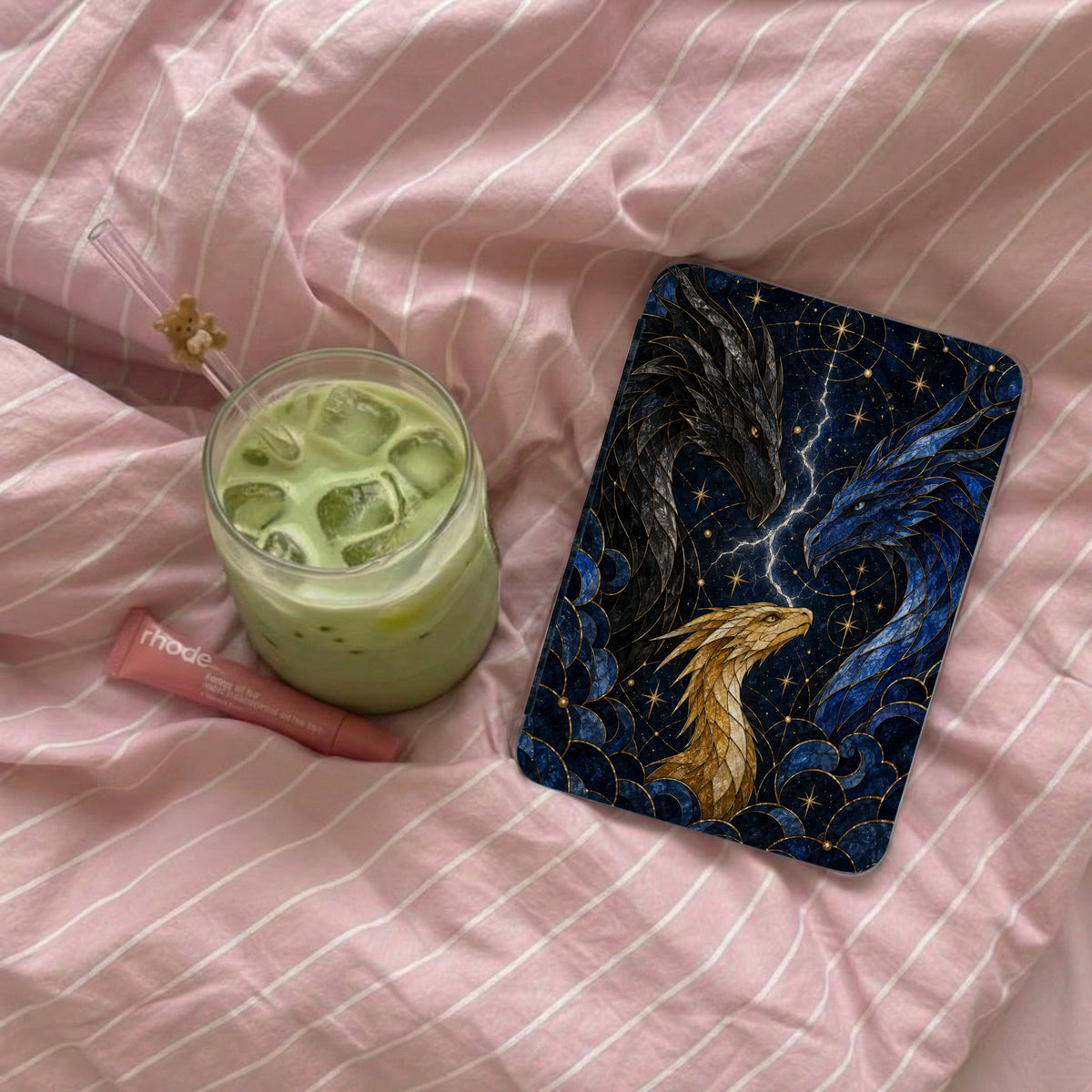Phone case with three dragons, iced matcha latte, and lip balm on pink striped bedding.