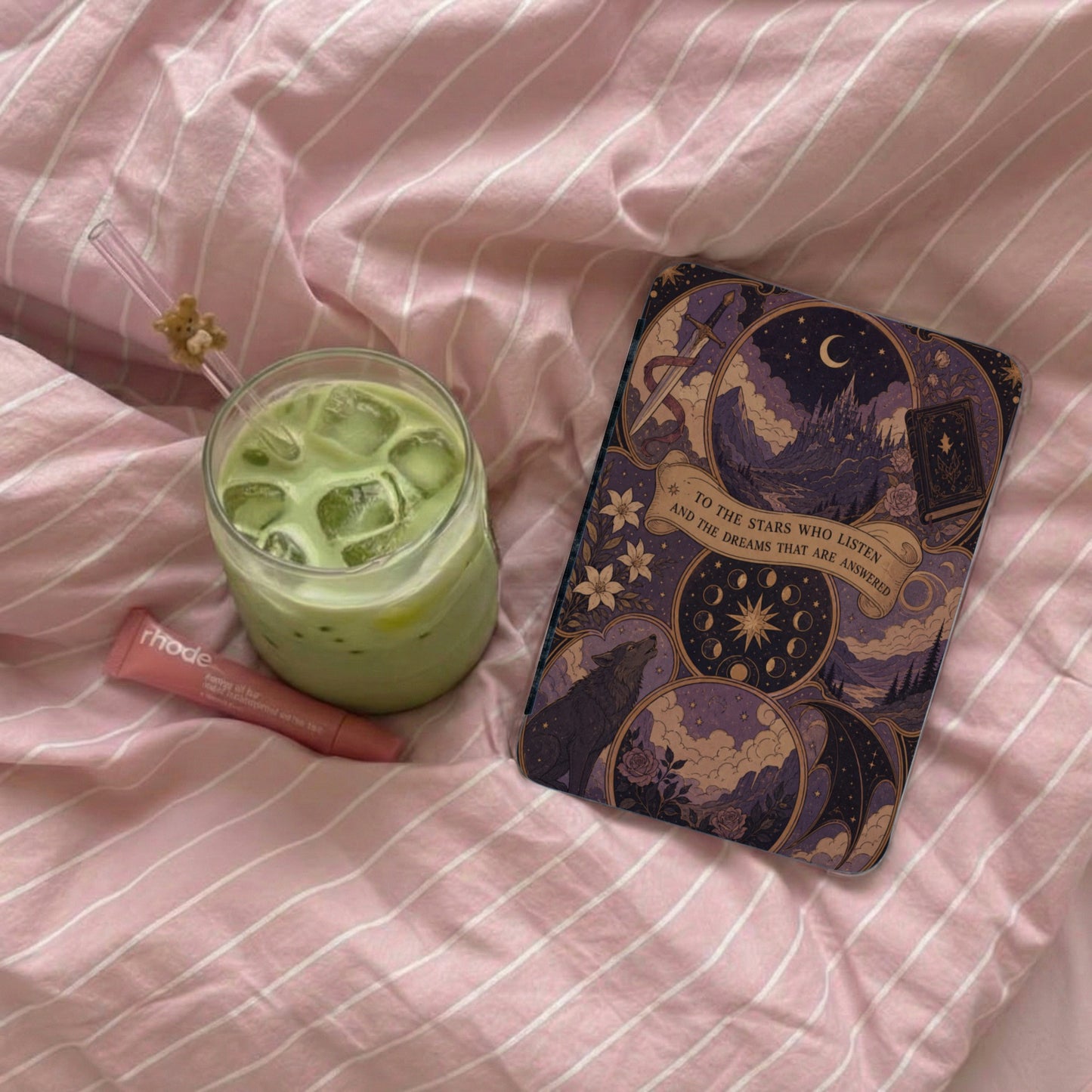 Phone case with celestial, wolf, and castle design next to iced matcha on pink striped bedding.
