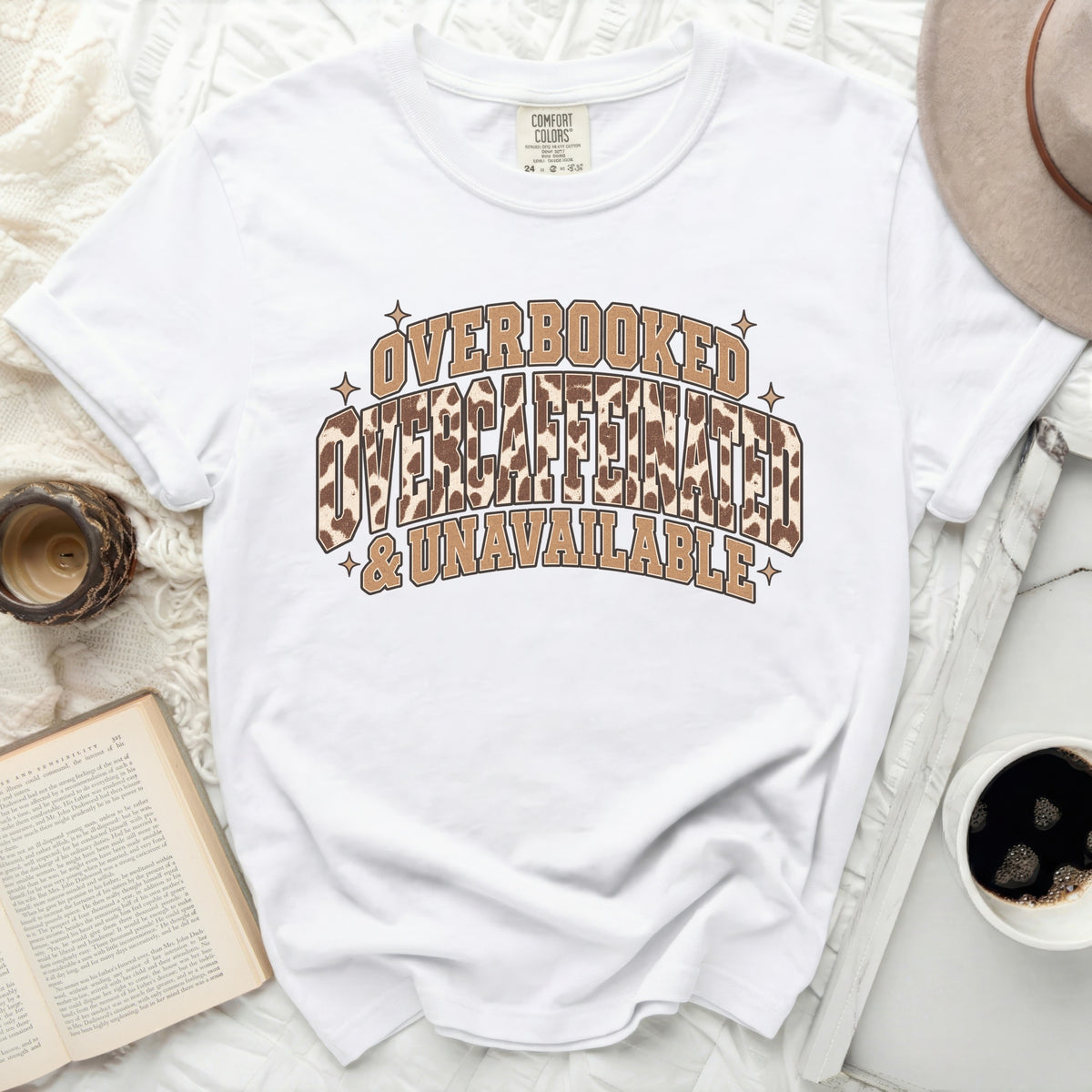 White t-shirt with text "OVERBOOKED OVERCAFFEINATED & UNAVAILABLE" in brown lettering.