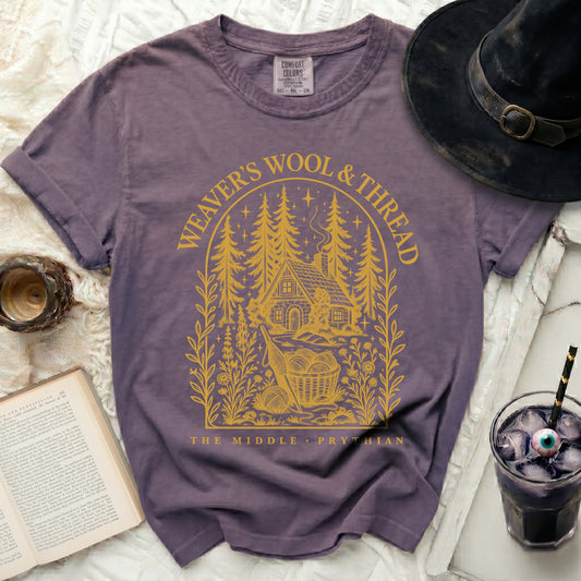 Purple t-shirt featuring a yellow graphic of a cabin in the woods with text reading Weaver's Wool & Thread.