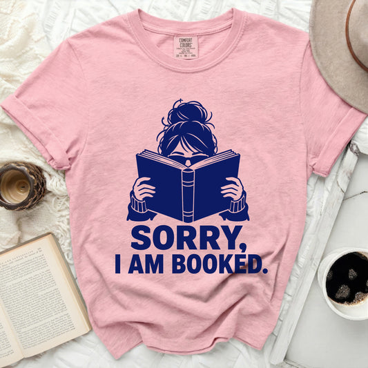 Pink t-shirt with dark blue graphic of a woman reading and text "SORRY, I AM BOOKED."