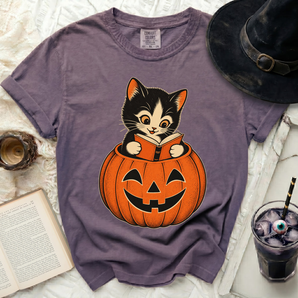 Kitten reading a book in a jack-o'-lantern on a purple t-shirt.