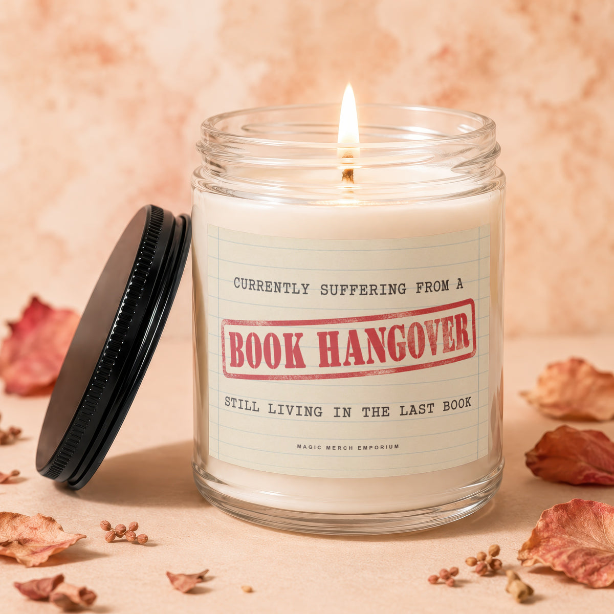 Candle with "Book Hangover" label and burning wick, surrounded by dried leaves.
