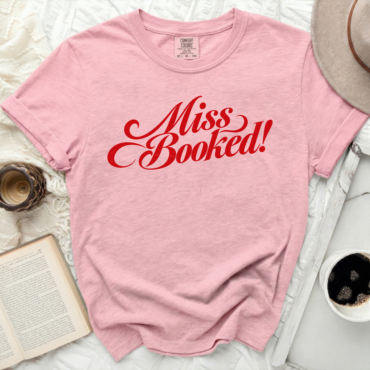 Pink t-shirt with red "Miss Booked!" graphic, flat lay with book and hat.