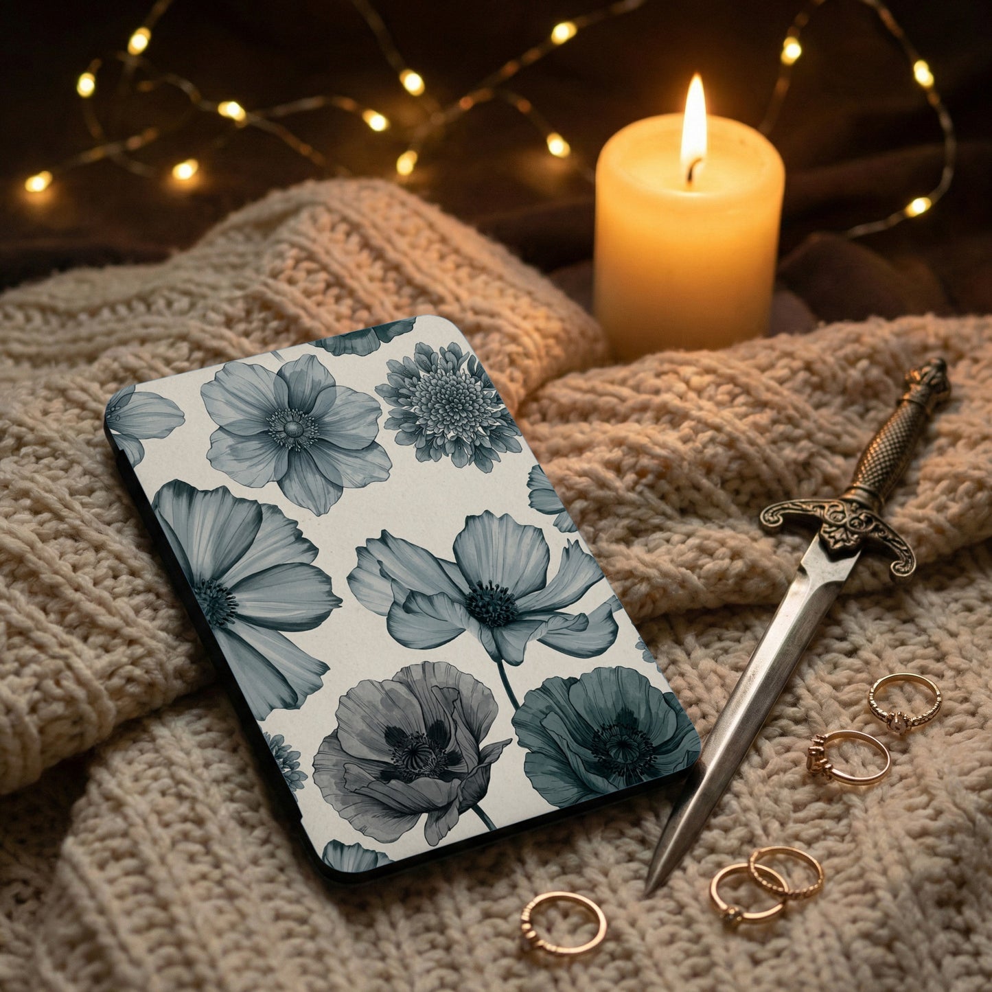 Case with blue floral pattern on beige knit fabric, near a lit candle and rings.