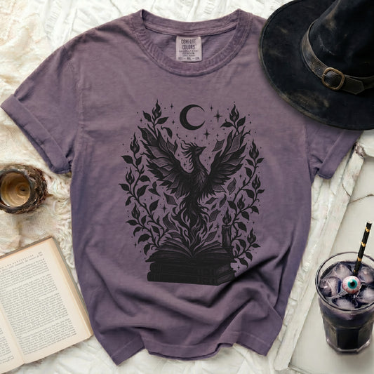 A purple crewneck t-shirt featuring a black graphic of a phoenix rising from books, surrounded by foliage and a crescent moon.