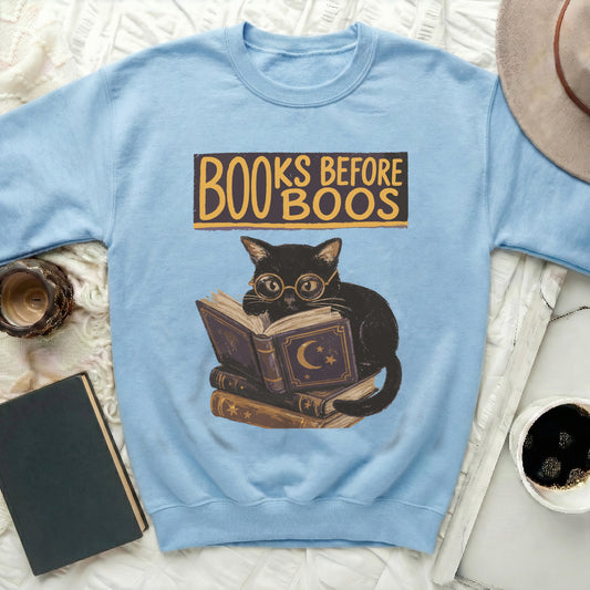 Light blue sweatshirt with graphic of black cat reading books, text "Books Before Boos".