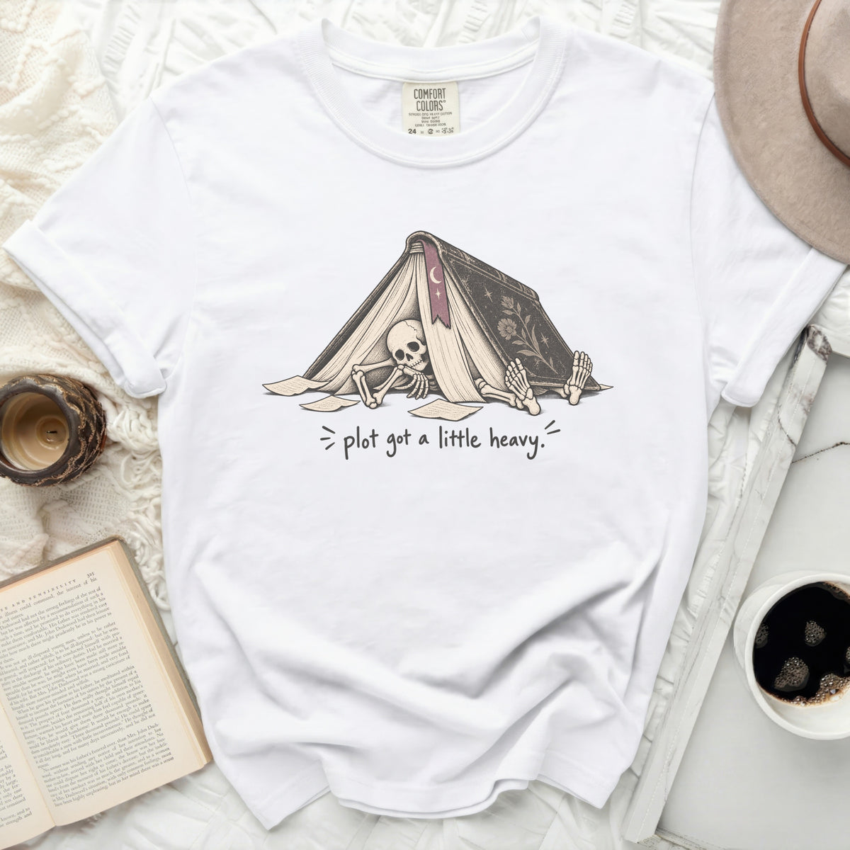 White t-shirt with graphic of skeleton under a book and text "plot got a little heavy."