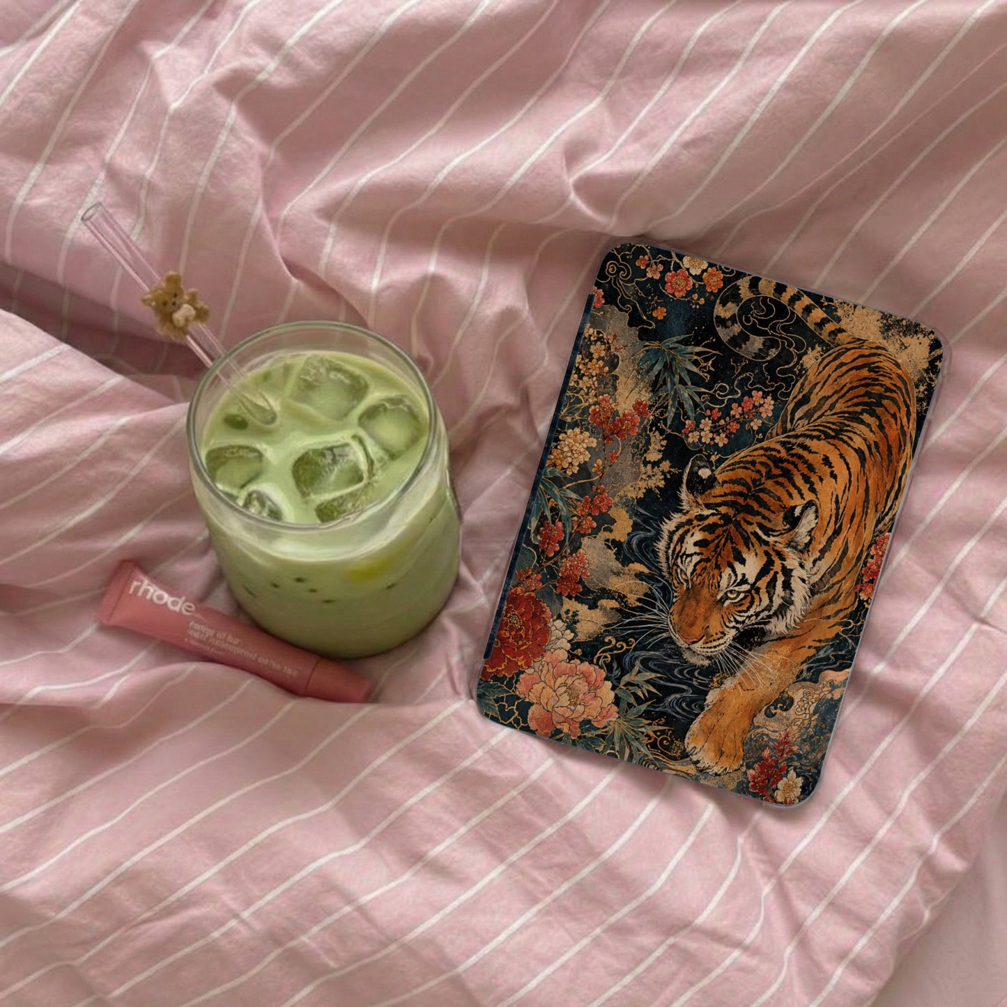 Phone case with tiger and floral design next to iced matcha drink on pink striped fabric.