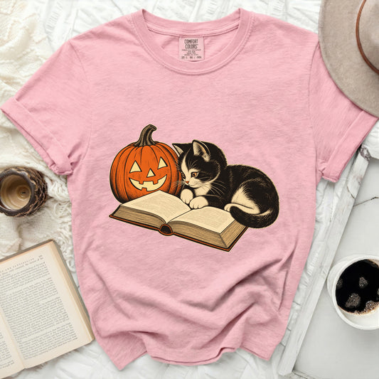 Pink t-shirt with a black and white kitten reading a book next to a jack-o'-lantern.