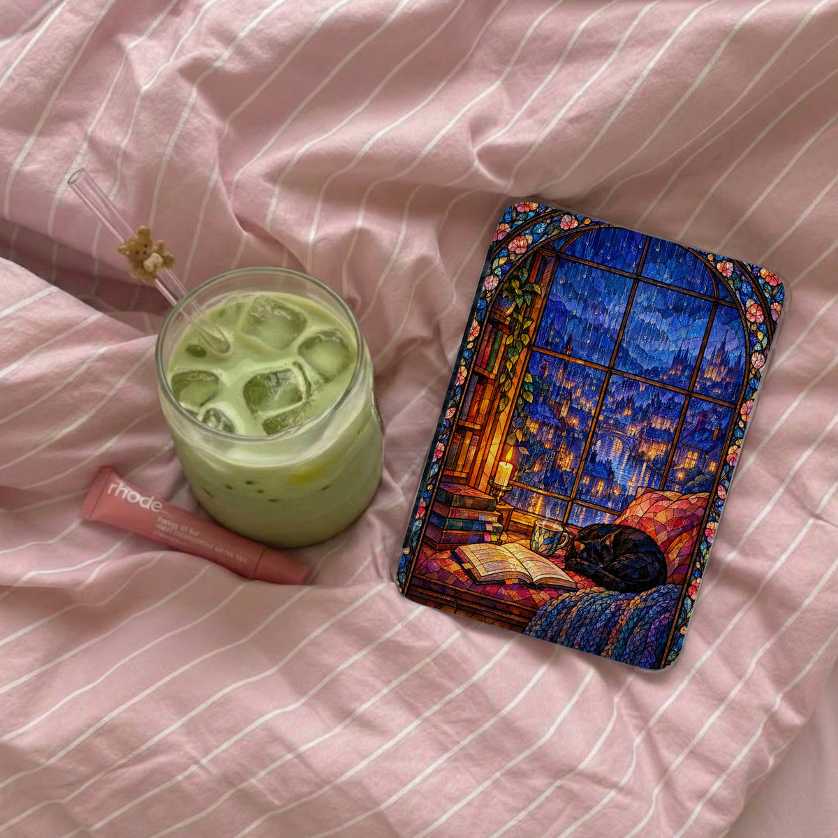 A Kindle in a stained-glass style case lies on a pink striped bed next to an iced matcha latte and lip balm.