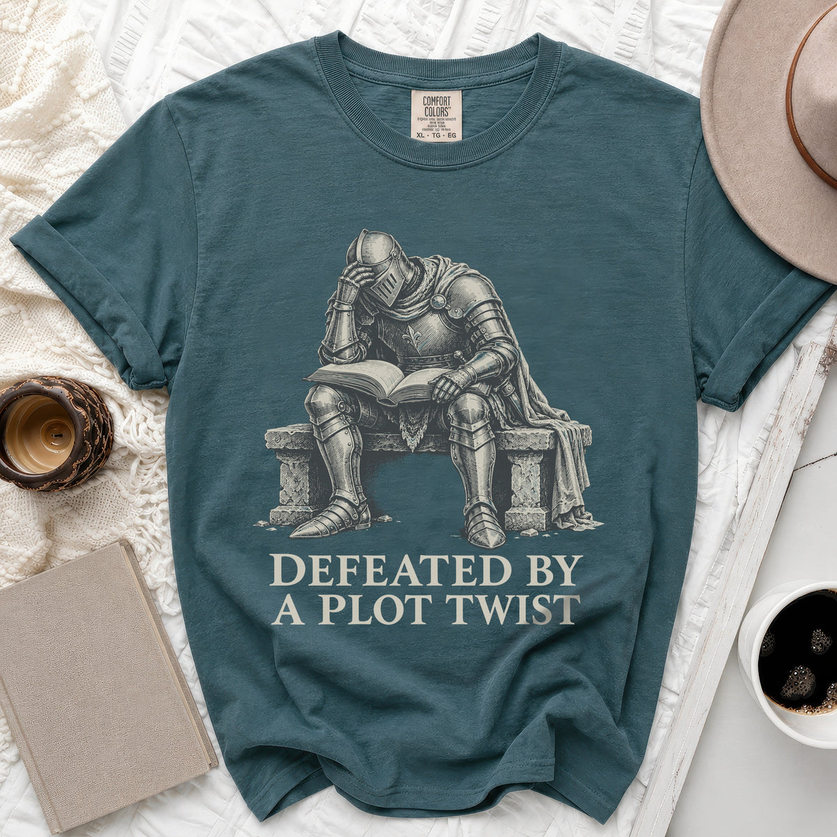 Teal t-shirt with a knight reading a book with text "Defeated by a Plot Twist".