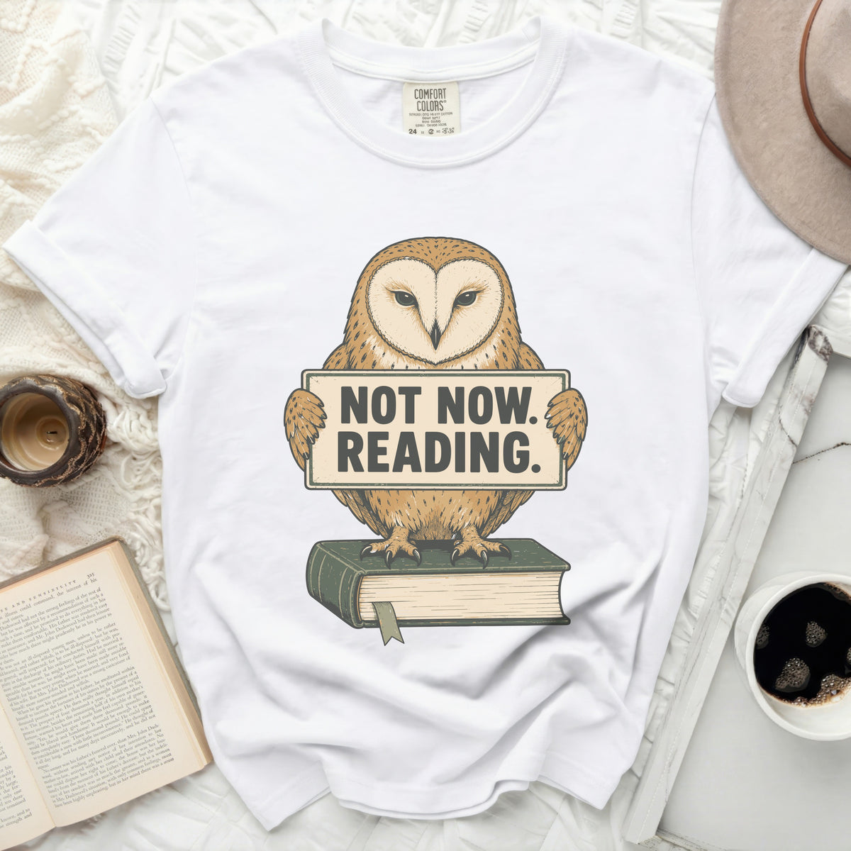 White t-shirt with barn owl graphic holding "NOT NOW. READING." sign and sitting on a book.