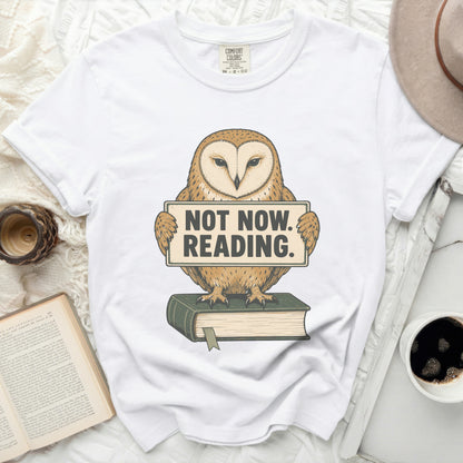 White t-shirt with barn owl graphic holding "NOT NOW. READING." sign and sitting on a book.