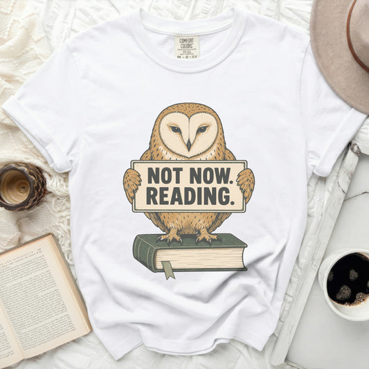 White t-shirt with barn owl graphic holding "NOT NOW. READING." sign and sitting on a book.