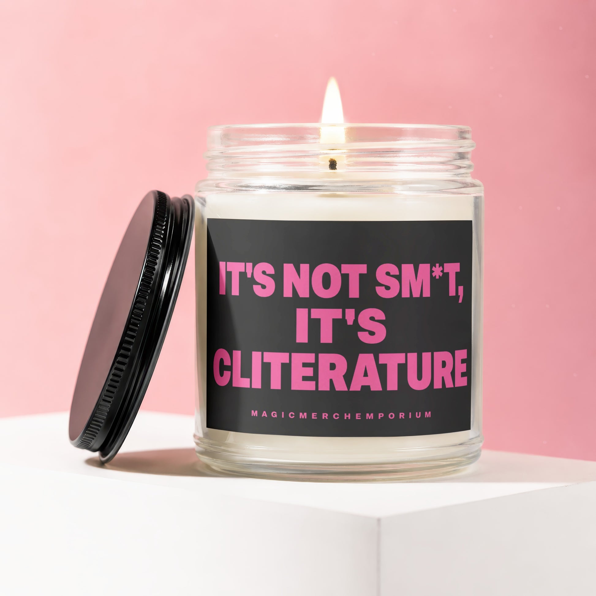 Glass candle with lit wick and black lid, reads "It's not s*m*t, it's cliterature".