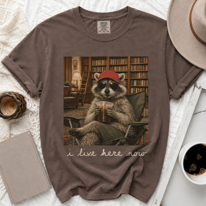 T-shirt with a raccoon in sunglasses and a red hat drinking an iced coffee with the text "i live here now".