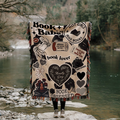 A person is standing on a rock, holding a blanket with various book-related designs and text, including phrases like "Book Babe", "Hot Girls Read Books", "book lover", "book observer", and "dark romance".
