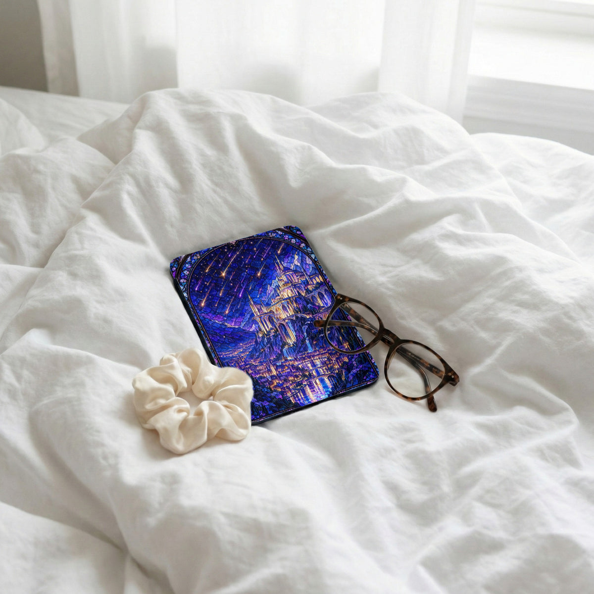 A Kindle reader with a stained-glass style castle cover rests on rumpled white bedding alongside tortoiseshell glasses and a cream scrunchie.