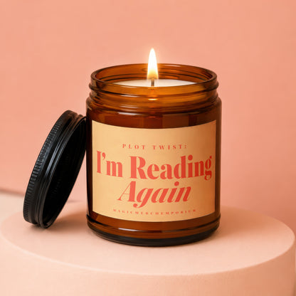 Amber candle with lit flame and black lid, featuring text "PLOT TWIST: I'm Reading Again".