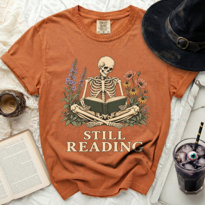 Orange t-shirt with skeleton reading surrounded by flowers and text "STILL READING."